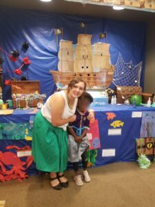 Taylor stands, bent over slightly, next to a child. behind them is a sea-themed wall decorated with pirate ships, coral, and other ocean-themed decorations. Taylor smiles at the camera with her arm around the child (whose face is blurred).