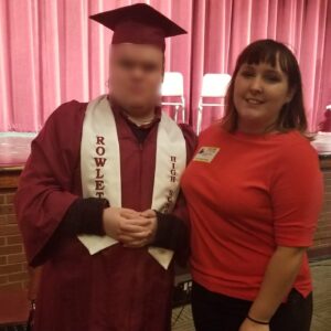 A photo of Taylor (right), next to her cousin, Billy (left). Bily's face is blurred for privacy. Billy wears a graduation cap and gown in a maroon color. The stole reads: "Rowlett High School." Taylor smiles, her arm intertwined with Billy.