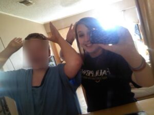 A mirror selfie of Taylor (right) next to her cousin, Billy (left). They both have their hands on their heads, fingers extended in a silly pose as if they're making moose "antlers" for themselves. Taylor smiles wide. Billy's face is blurred.