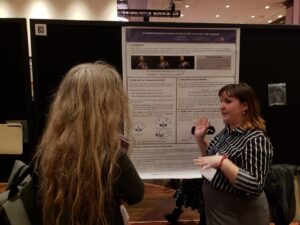 A photo of Taylor presenting a poster at a conference. She stands in front of the poster (which contains pictures and diagrams of sign language), speaking to another attendee (left). Her hands move as she talks.