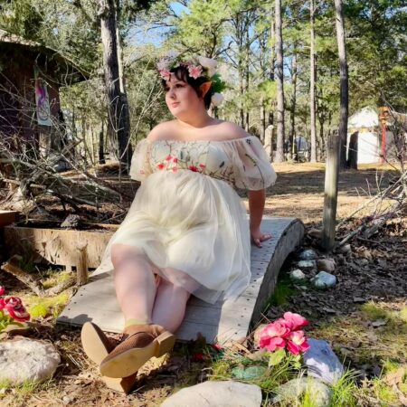 A picture of taylor in a fairy costume-- a dres with embroidered flowers, and a matching flower crown. She sits on a small wooden bridge, peering off into the distance. The woods are in the background.