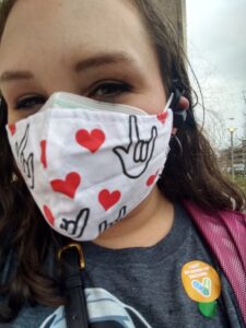 A close-up selfie of Taylor, wearing a face mask with a sign language and heart pattern on it. She appears to be smiling, and wears a sticker indicating she just received her COVID vaccine.