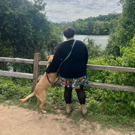 A shot of both Taylor and her dog, Cubby, from behind. They stand in front of a fence that overlooks a small river. There are abundant plants around. Taylor has her hand on Cubby, who rests his paws against the top of the fence.