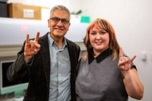 Taylor (right), stands next to her advisor (left), Dr. David Quinto-Pozos. Taylor is a white woman with brown and blonde hair, wearing a formal gray dress. David has tan/brown skin and short gray hair, wearing glasses and a button-up shirt with a suit jacket. They both smile at the camera, holding up a "University of Texas" hand sign.