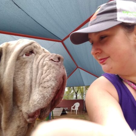 A selfie of Taylor with a massive, grey, dog (a mastiff mix). Taylor and the dog look at each other, Taylor smiling softly at the dog. She wears a ballcap and gear for walking dogs.