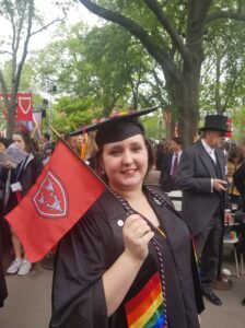 A picture of Taylor at graduation. She stands outdoors, wearing her graduation cap and gown. She wears a red and black cord (signifying first-generation status), a rainbow stole, and waves a small flag with the logo for Dunster House on it (three moose heads on a shield).