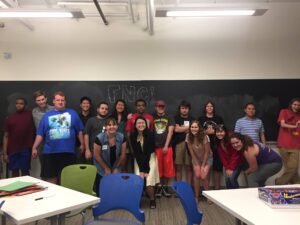 A group photo of Friday Night Club attendees. People of various ages stand in front of a blackboard, smiling at the camera and posing.