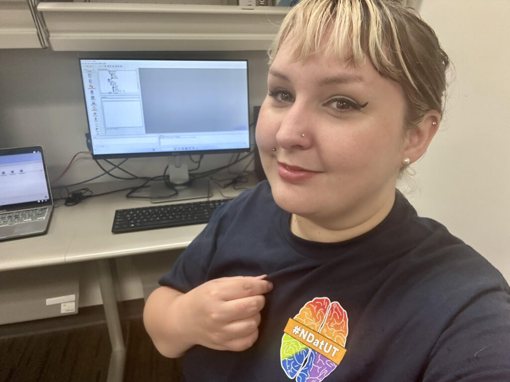 A selfie of Taylor in an office/laboratory setting. She has split-dye hair (half brown, half blonde), and smiles at the camera. Behind her is a lab computer with software running. Taylor points to her own t-shirt, which has a cartoon of a rainbow brain on it with the label "Neurodivergent at UT."