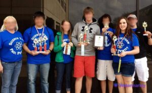 A photo of Taylor's science team and her coach. All of the faces besides Taylor's are blurred; everyone looks into the camera, holding various medals and trophies. The t-shirts read: "Pewitt Science Team."