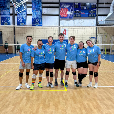 A group photo of Taylor and her volleyball team. Seven players stand on the court facing the camera, smiling with their arms around each other. Taylor is the second to last person from the right. The volleyball gym is behind the team.